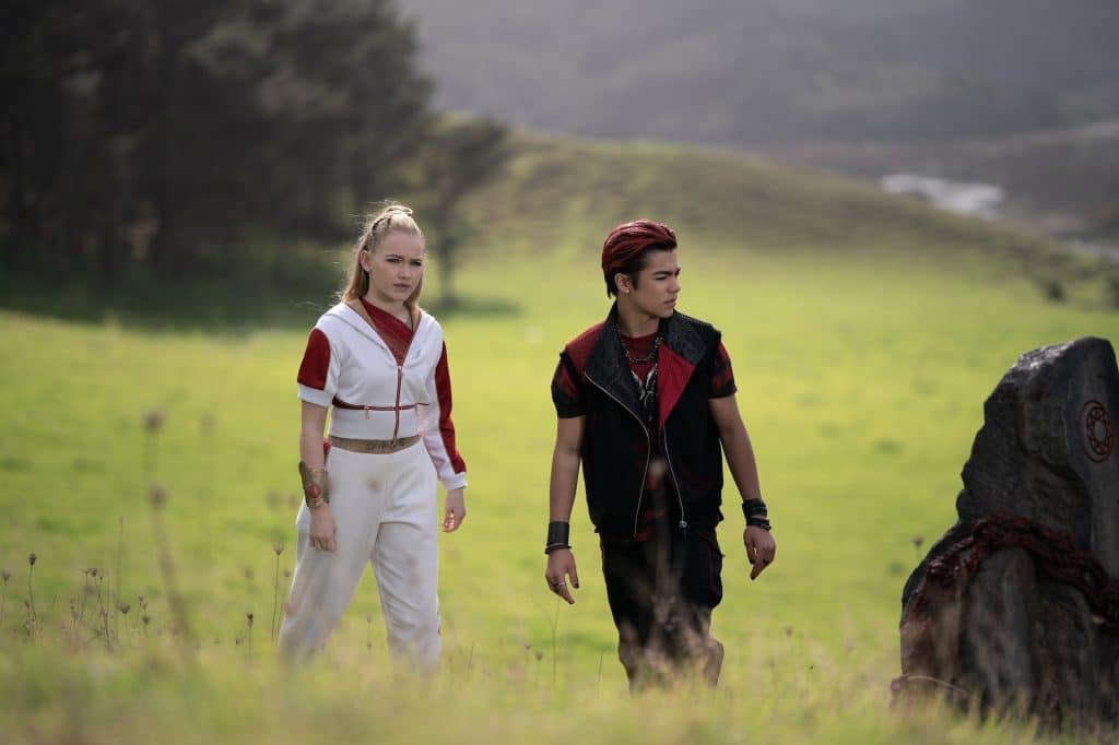 Two young people in sporty, futuristic outfits stand in a grassy field near a large stone with red markings, as if protecting secrets of the sea. Trees and hills are visible in the background under soft, natural light, lending an air of mystery reminiscent of Zombies 5.