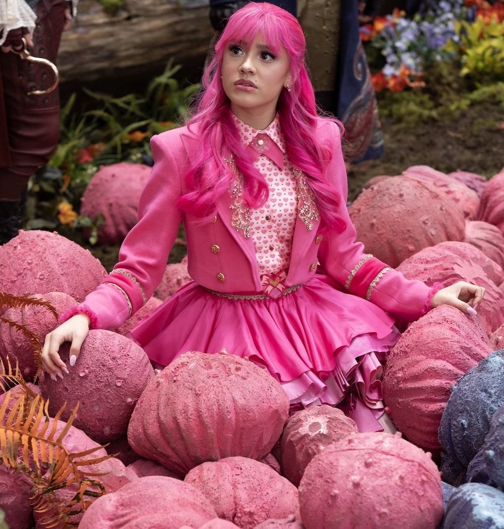 A woman with bright pink hair and a detailed pink outfit sits among large, textured pink boulders in an outdoor, whimsical setting where worlds collide, looking up with a curious expression.