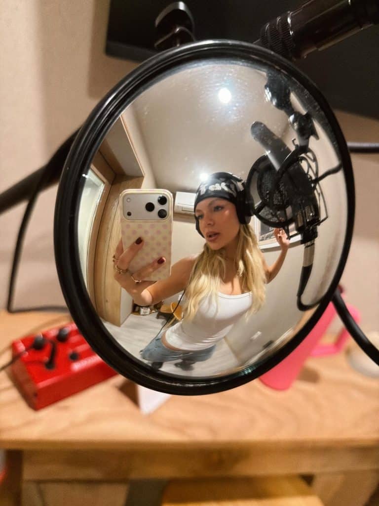 A woman wearing headphones and a beanie takes a selfie in a recording studio, reflected in the pop filter of a microphone—capturing a candid moment that channels Pressley Hosbach’s creative energy.