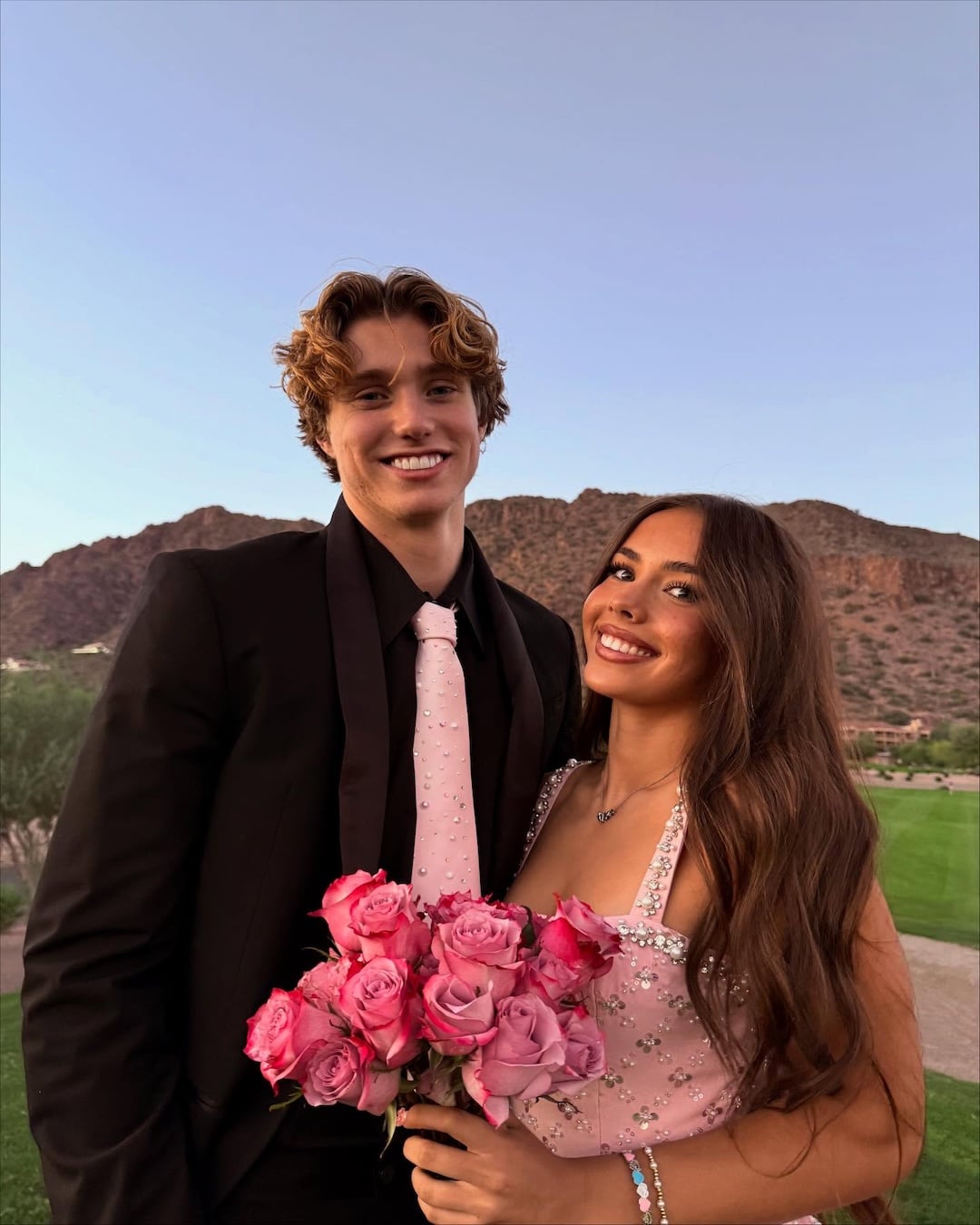 A young couple dressed in formal attire stands outdoors, smiling. The young woman holds a bouquet of pink roses and wears a pink dress, while the young man wears a black suit with a pink tie—the perfect summer crush moment against a mountain backdrop.