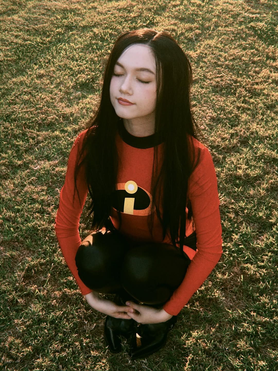 A person with long dark hair, eyes closed, is crouching on grass while wearing a red superhero costume top with a black “i” logo, similar to Violet Parr from The Incredibles. Sunlight softly illuminates their face.
