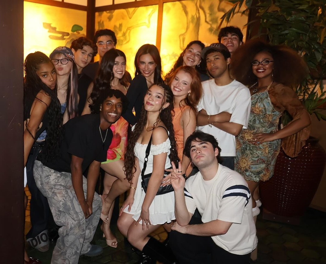A group of teens and young adults pose together, smiling at the camera in front of a brightly lit backdrop—unbothered by a recent party blunder involving someone's little bro—some standing and some kneeling, all dressed in casual and trendy outfits.
