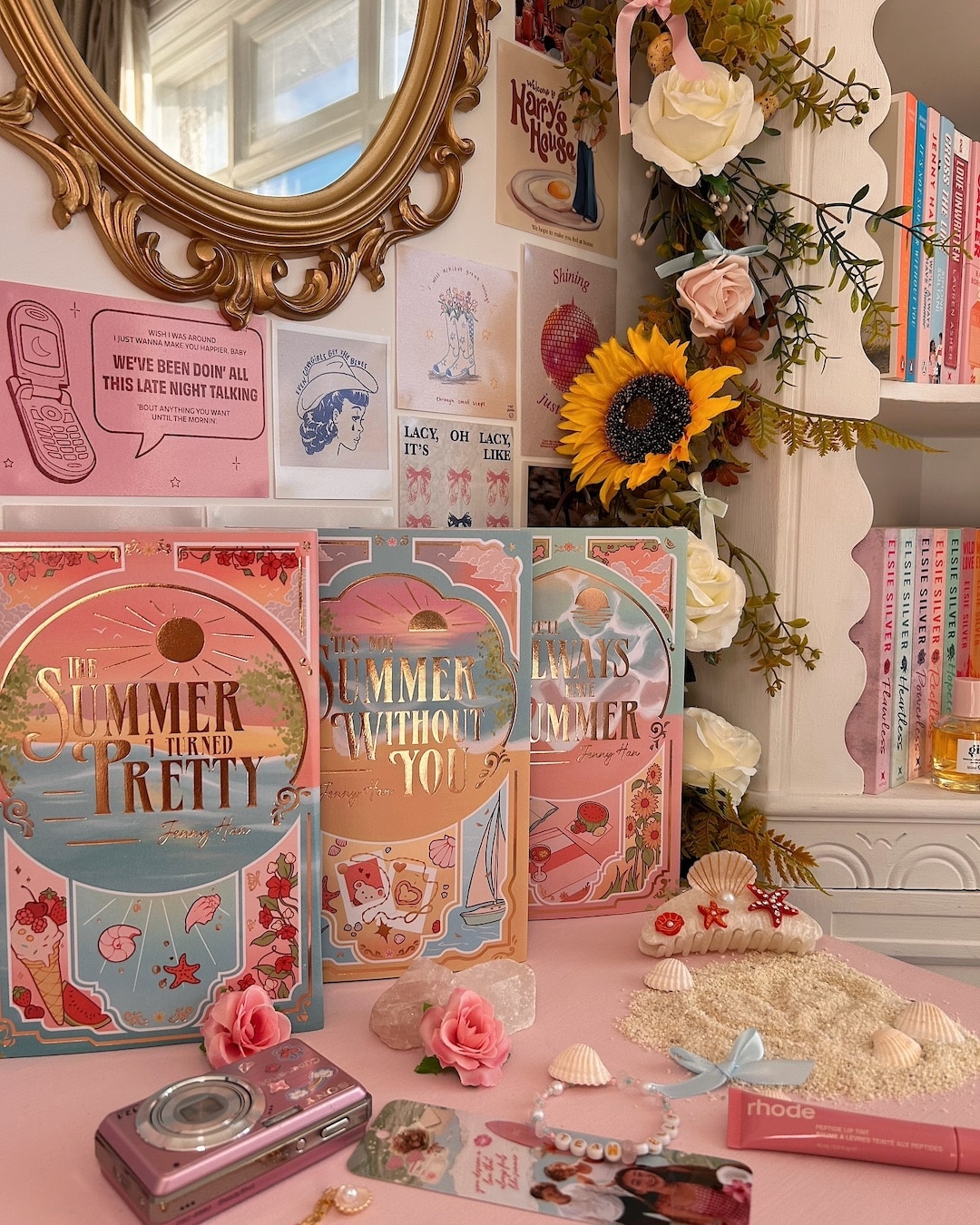 A pastel-themed book display features TSITP (The Summer I Turned Pretty) series, roses, seashells, a digital camera, a sunflower, and a mirror. Books and postcards decorate the cozy, sunlit space.
