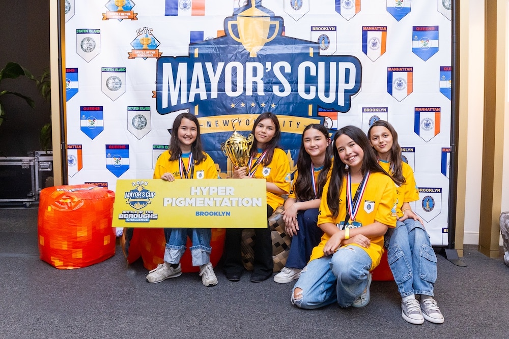 Five young women in yellow shirts sit and smile, radiating girlpower as they hold a trophy and a sign that reads Hyper Pigmentation, Brooklyn, in front of a Mayor’s Cup backdrop decorated with various borough logos. Their achievement has placed them at the top of the leaderboard.