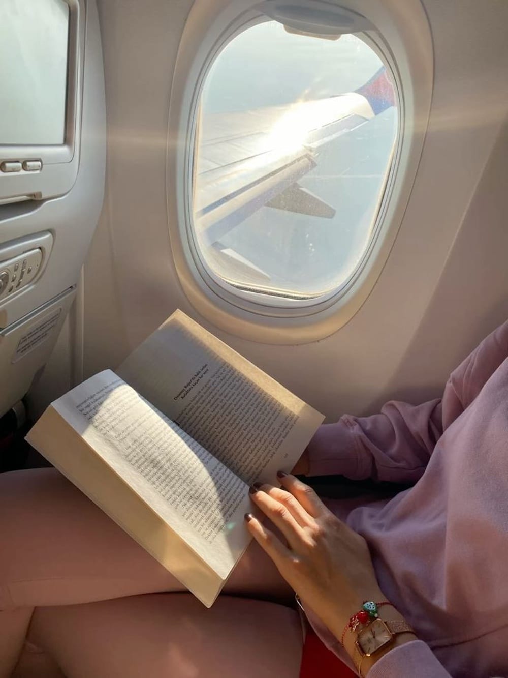 A person in pink clothes reads a summer read by the window seat of an airplane. Sunlight streams through the window, illuminating the book and part of the airplane wing outside, setting the perfect scene for a dream vacation.