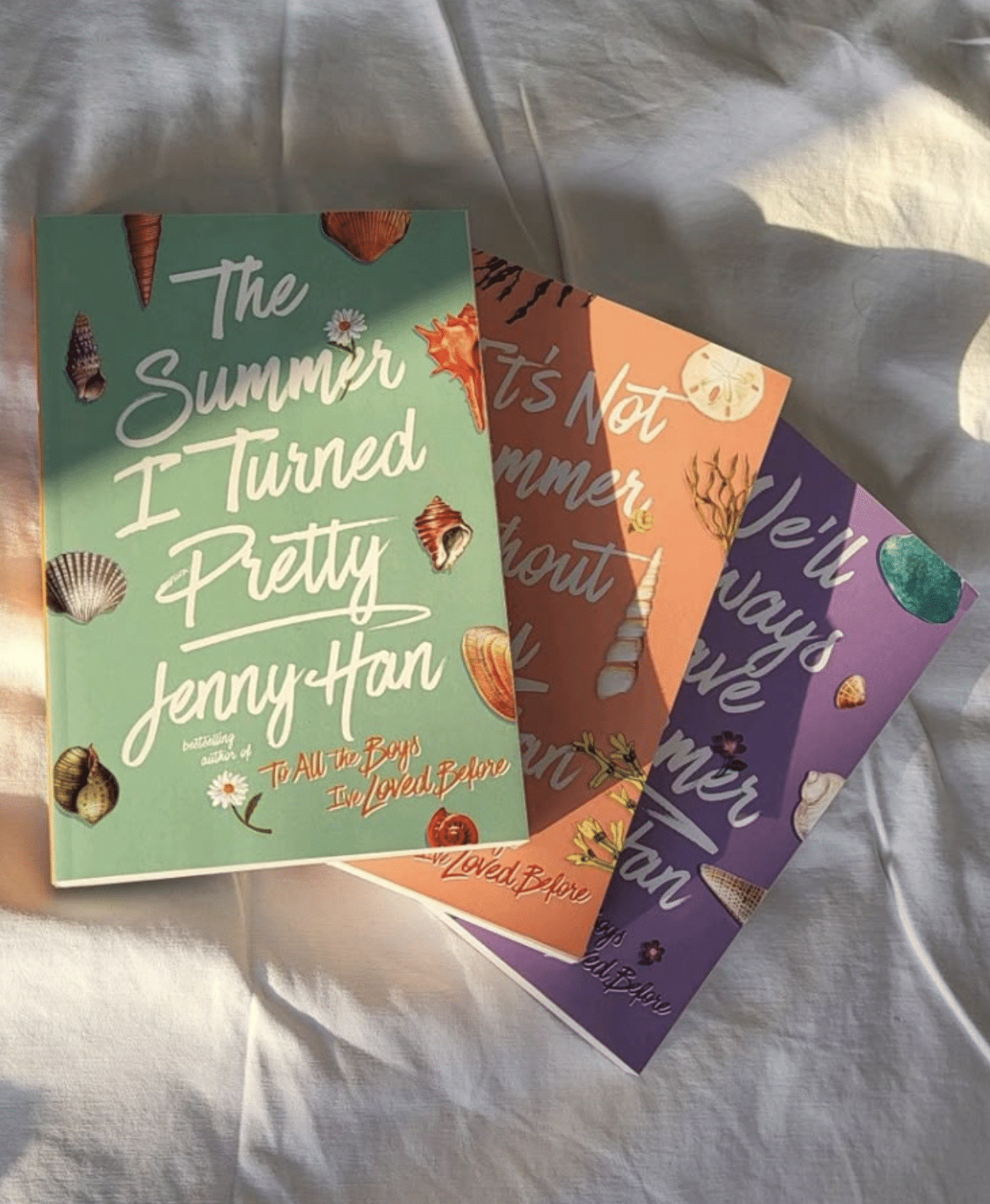 Three pastel-colored books from Jenny Han’s TSITP (The Summer I Turned Pretty) series are laid out on a sunlit white bedsheet, their covers featuring delicate seashell illustrations—a dreamy addition to any TBR and perfect for summer book recommendations.
