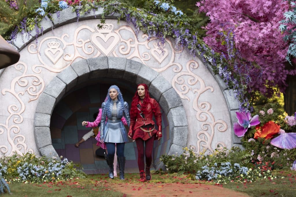 Two women dressed in colorful fantasy costumes stand in front of a round stone doorway surrounded by flowers, heart designs, and vibrant pink and purple foliage, suggesting a magical or fairytale setting inspired by the descendants of a wicked wonderland.
