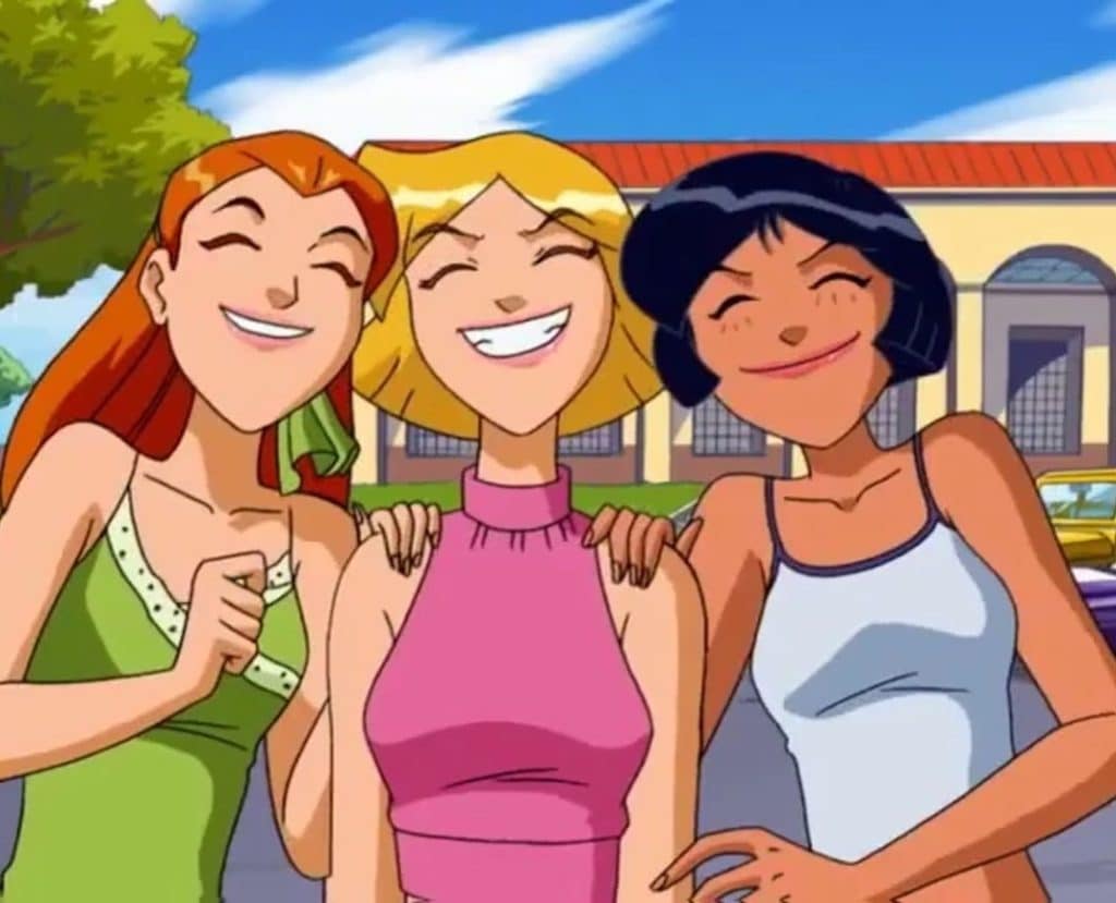 Three animated girls smile happily together outdoors, with their arms around each other. There are buildings and trees in the background under a bright blue sky.