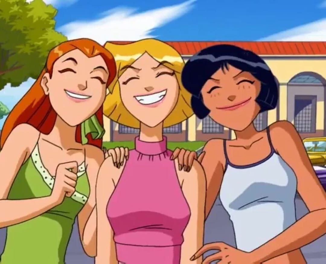 Three animated girls smile happily together outdoors, with their arms around each other. There are buildings and trees in the background under a bright blue sky.