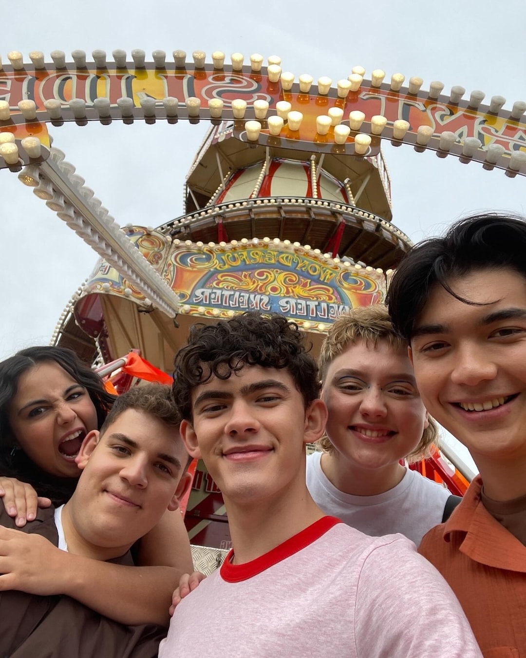 Five smiling teenagers take a selfie in front of a colorful amusement park ride with bright lights and a vintage design.