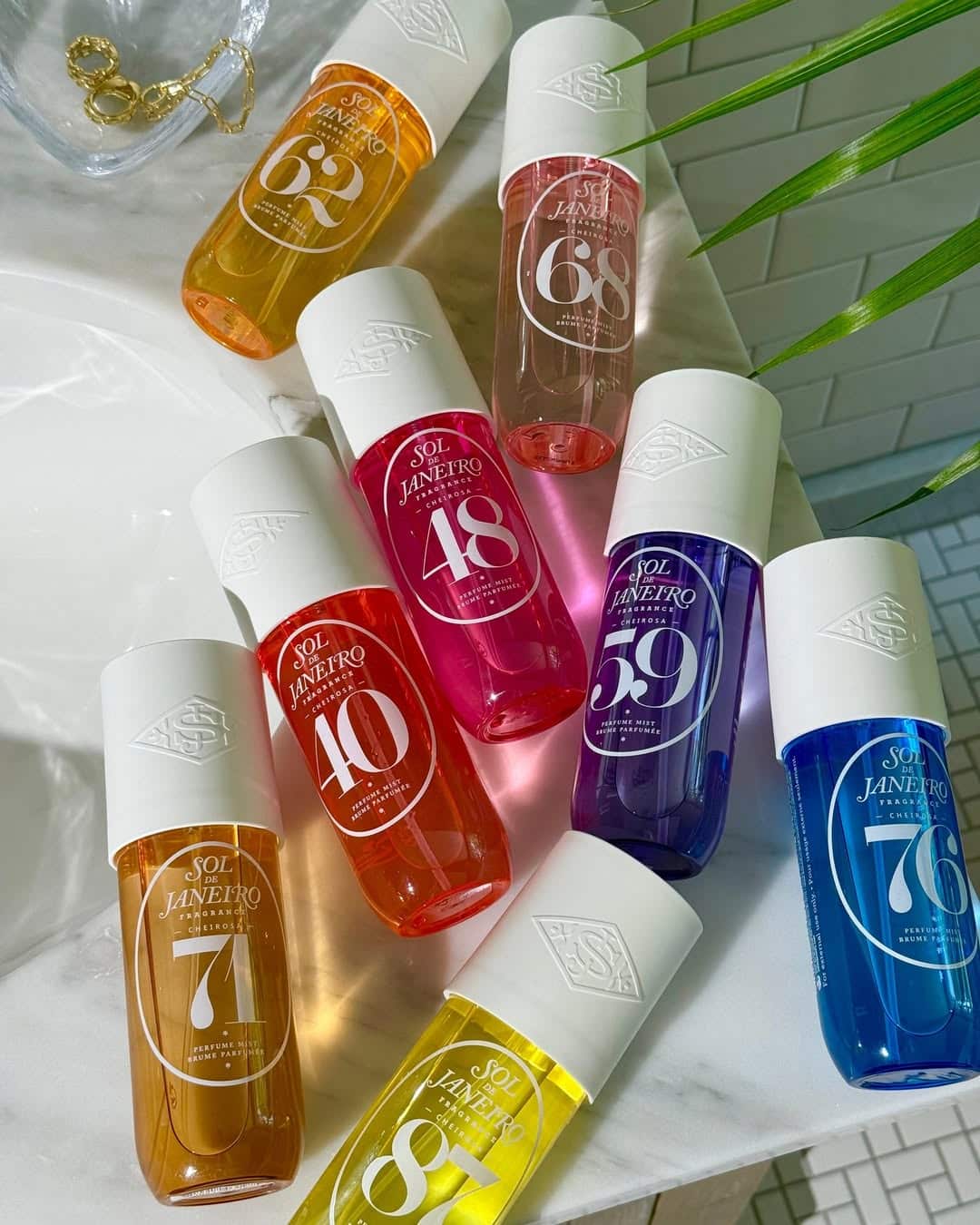 Eight colorful Sol de Janeiro body mist bottles with white caps are arranged on a white surface next to a glass dish and green plant leaves. Each bottle features a different number and vibrant hue, making it easy to find your perfect scent or explore a fragrance guide for wearing perfume every day.