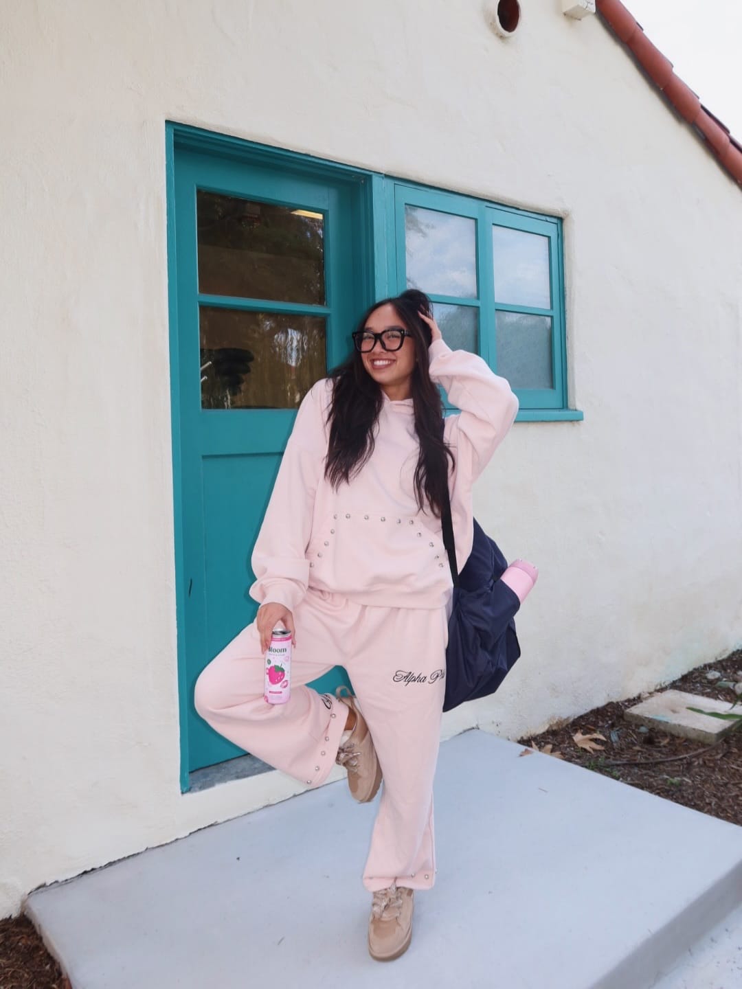 Woman in light pink sweatsuit and glasses stands smiling in front of a teal door, holding a drink and a black bag, with one foot raised playfully and a yoga mat tucked under her arm—channeling the positive energy of beloved book characters ready to take on new life lessons this back-to-school season.