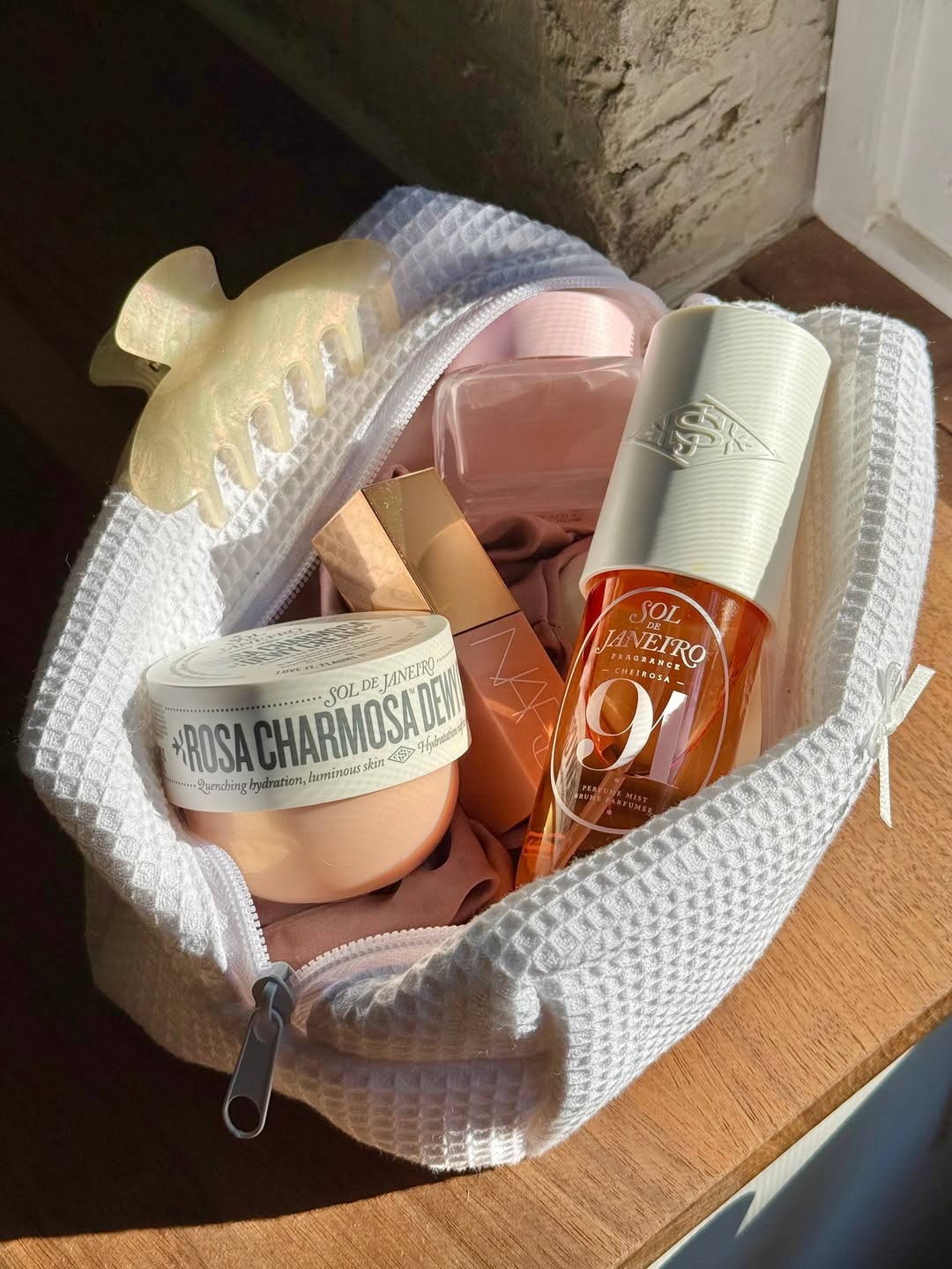 A white toiletry bag holds a hair clip, Sol de Janeiro Rosa Charmosa Dew cream, a Sol de Janeiro 9 perfume mist—the perfect scent for wearing all day—a peach bottle, and a nude lipstick, all bathed in sunlight by a window.