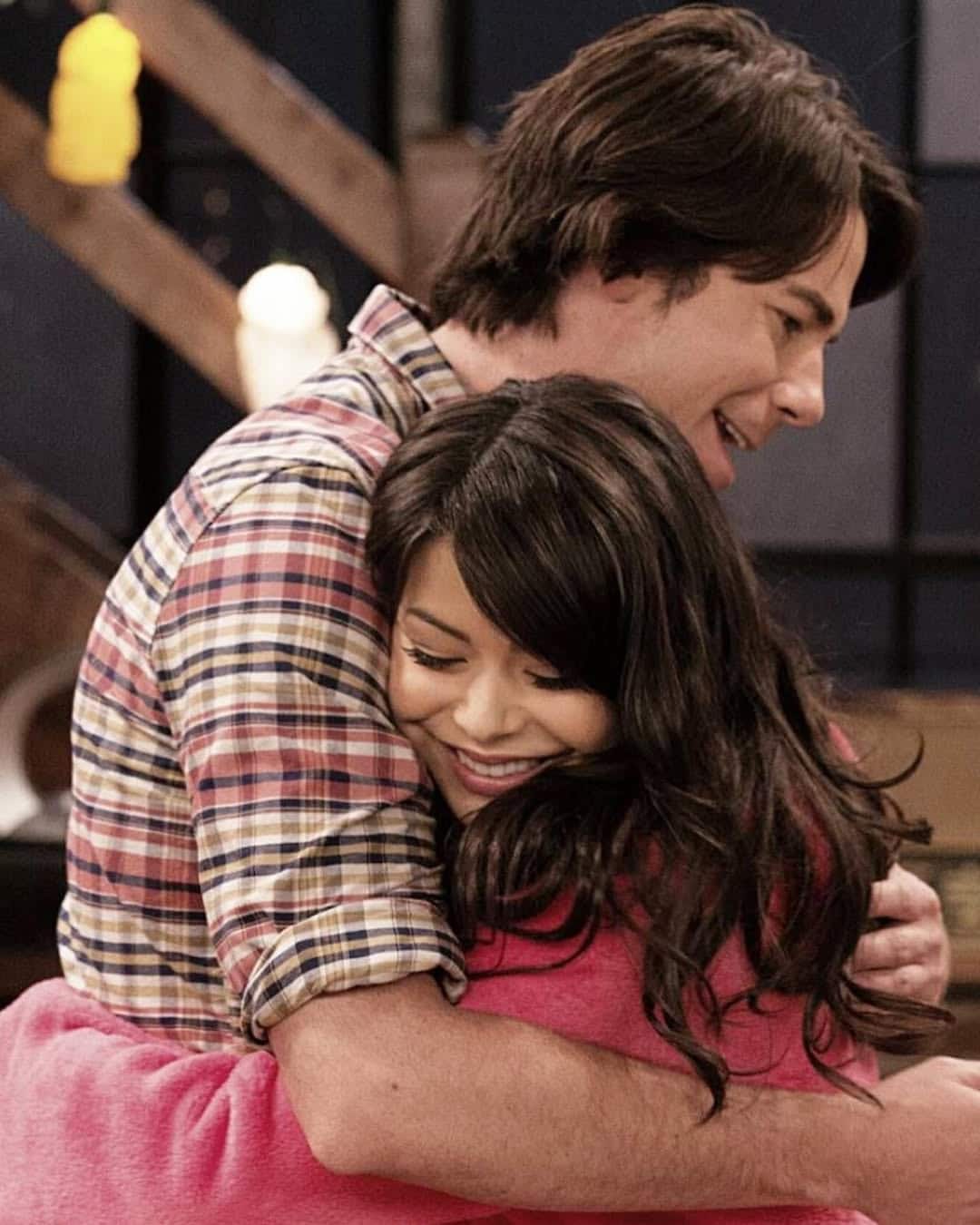 Two people warmly hug each other and smile. The person facing the camera has long dark hair and is wrapped in a bright pink blanket, while the other wears a plaid shirt. The background features wooden stairs and soft lighting.