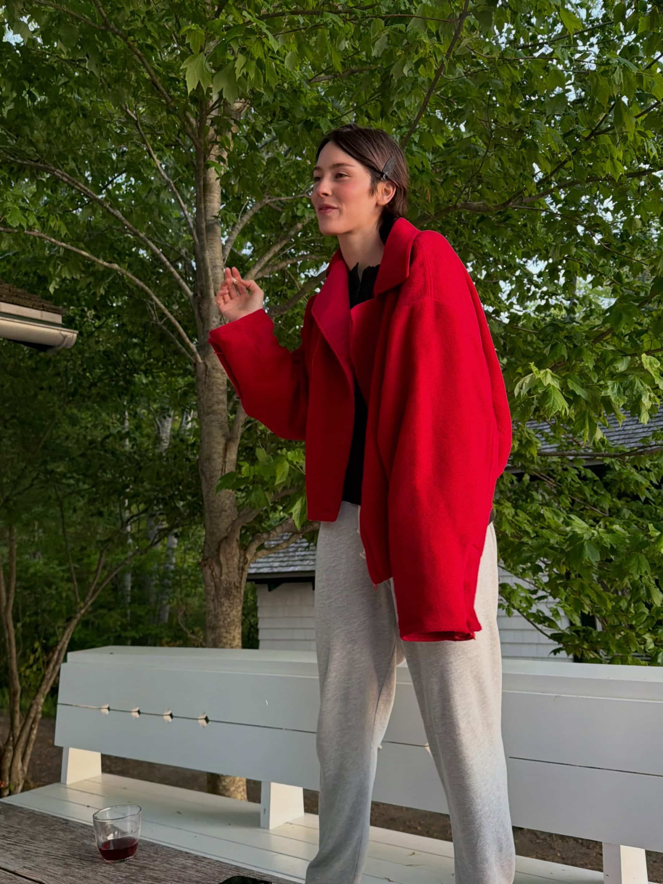 A person in a red jacket and gray sweatpants stands on a white bench outdoors, surrounded by green trees. A glass with a dark drink sits on the bench, perhaps enjoyed while listening to Gracie Abrams’ new album. The person appears relaxed and content.