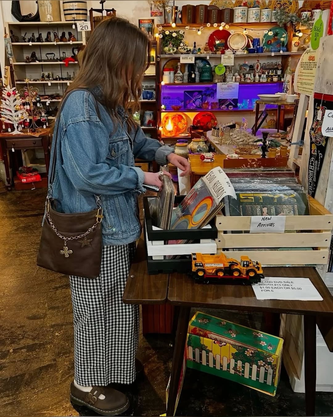 A woman, the unmistakable main character of her own summer adventure, wears a denim jacket and checkered pants as she browses vinyl records in a vintage shop filled with retro decor, nostalgic toys, and colorful lights—each corner promising the possibility of new side quests.
