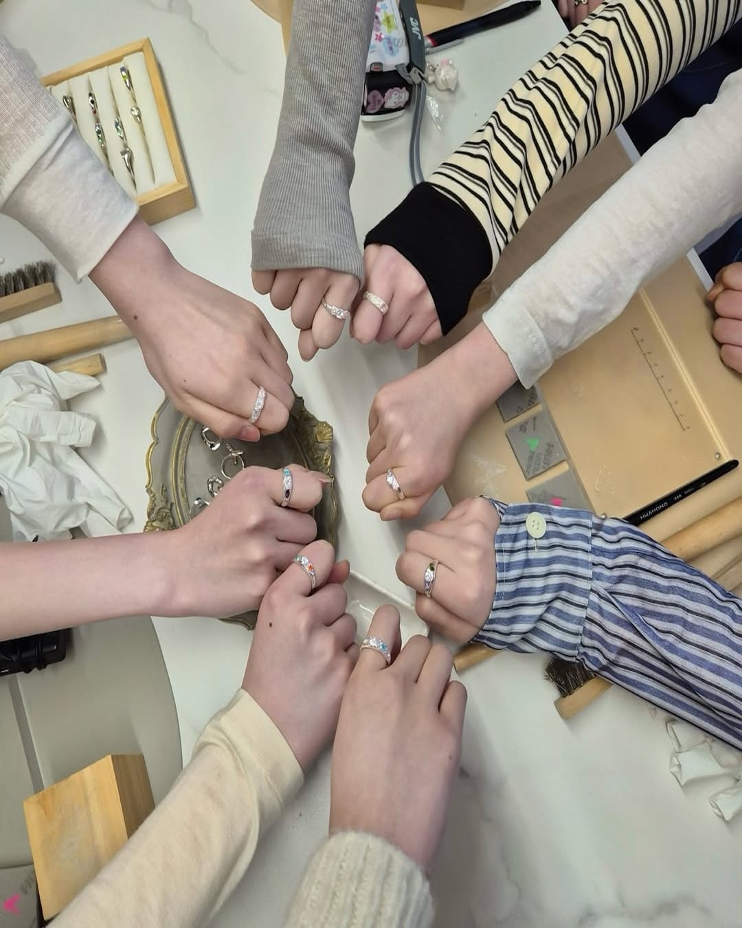Eight people sit around a table, each showing off rings on their left hands, fists meeting in the center. The table has notebooks, trays, tissue, brushes, and pens scattered around.