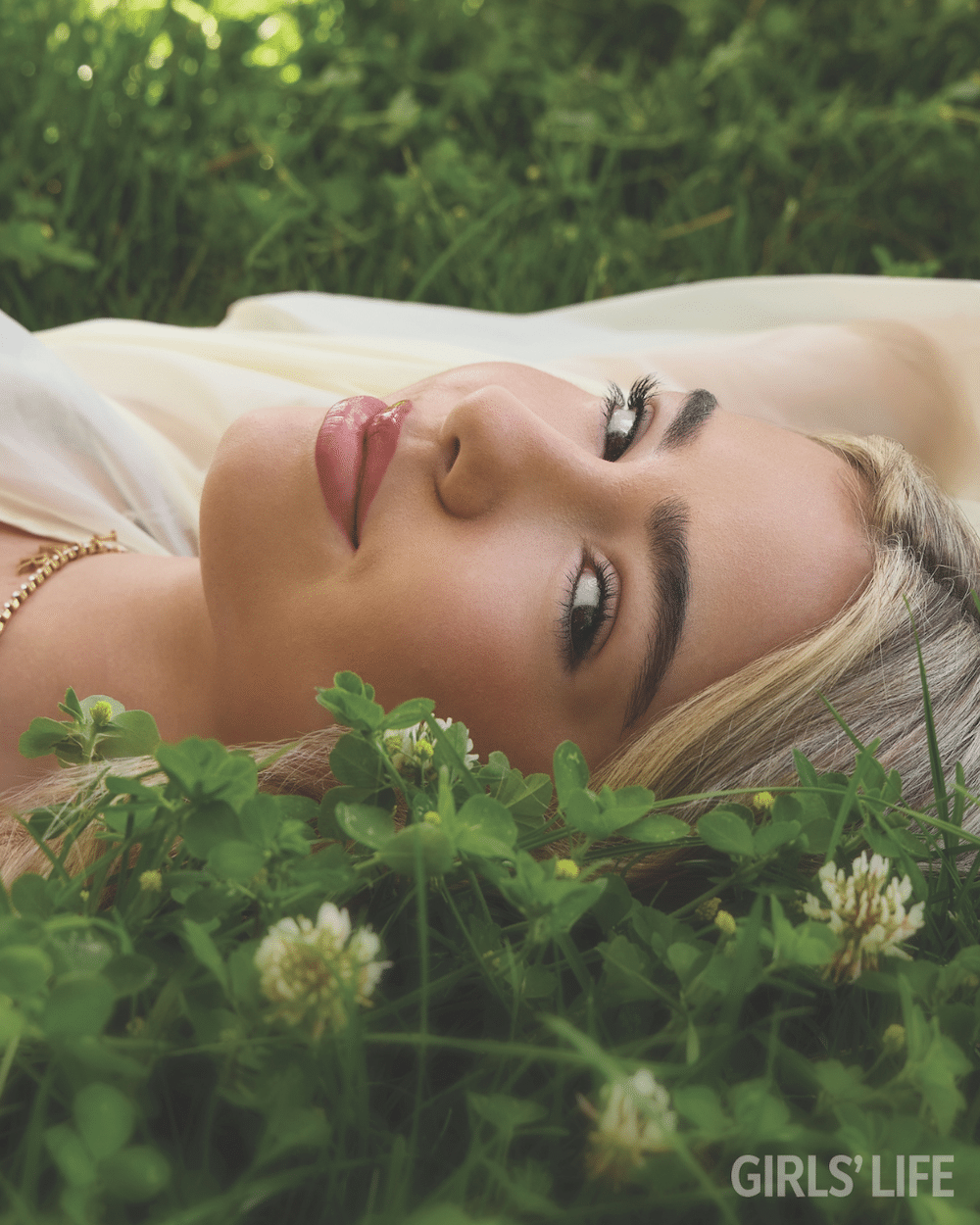 A young woman with blonde hair lies on green grass and clover, gazing at the camera. Sunlight highlights her face, capturing Embreigh Courtlyn’s serene expression. The GIRLS LIFE logo is visible in the lower right corner.