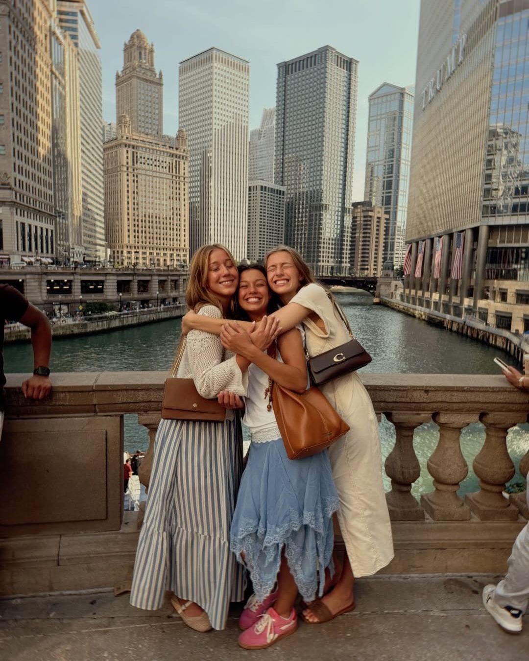 Three young women, feeling like the main characters of their own summer adventure, hug and smile on a bridge over a river, surrounded by tall city buildings on a sunny day.