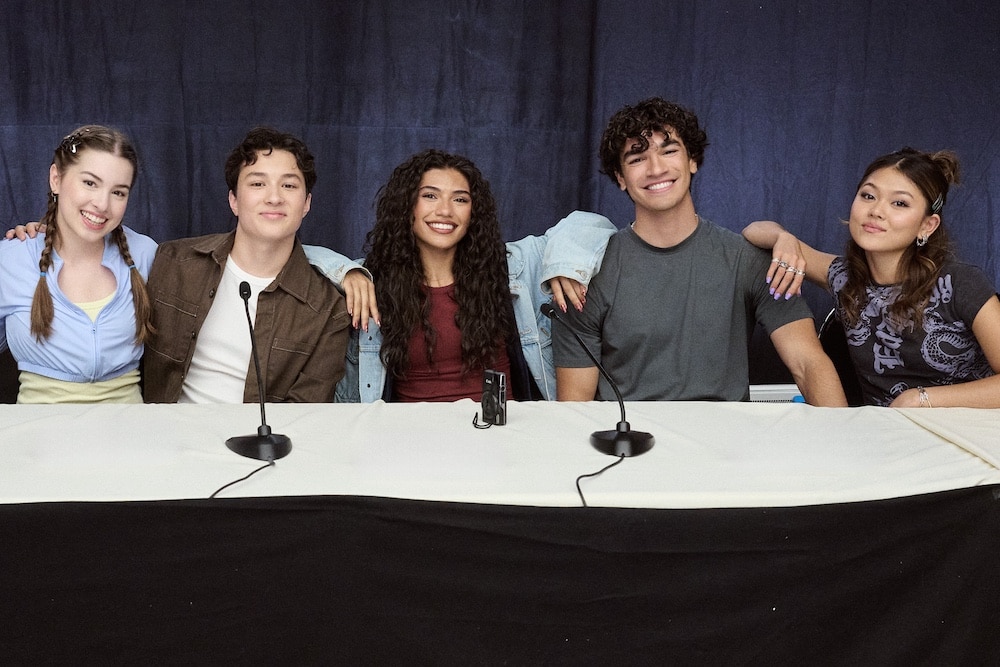 Five smiling teenagers, descendants of renowned local artists, sit behind a table with microphones, arms around each other, posing for a group photo against a plain dark backdrop.