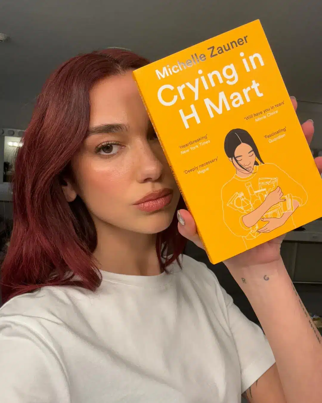 A person with red hair wearing a white shirt holds up the book Crying in H Mart by Michelle Zauner, partially covering their face—perhaps searching for their next read or taking inspiration from Gracie Abrams while waiting for her new album.