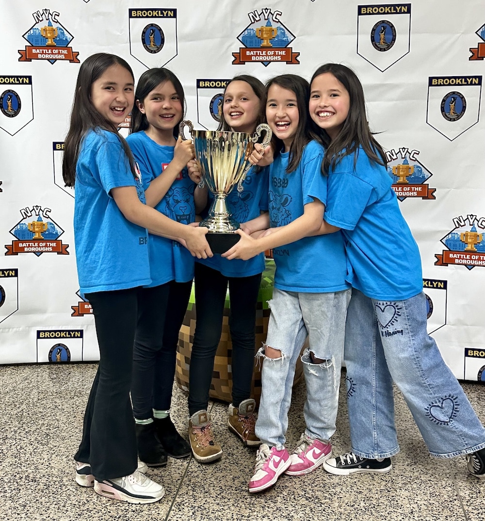 Five smiling children in blue shirts hold a large trophy together, celebrating their Minecraft victories as they stand in front of a Battle of the Boroughs NYC Brooklyn banner.