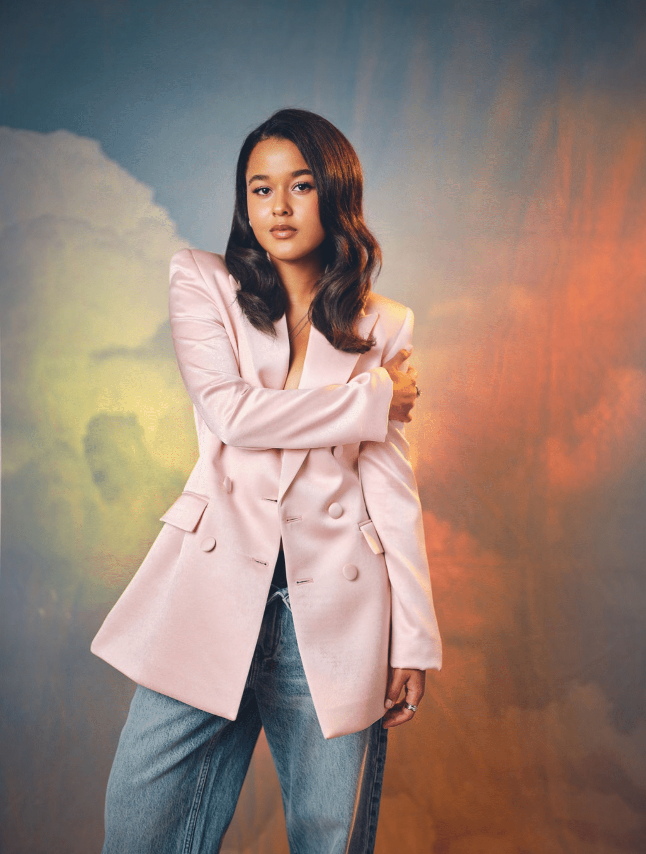 A woman with wavy dark hair wearing a light pink blazer and jeans stands with one arm crossed in front of a cloudy, softly lit background, reminiscent of the poised elegance often seen in Jordan Alexa Davis’s portraits.