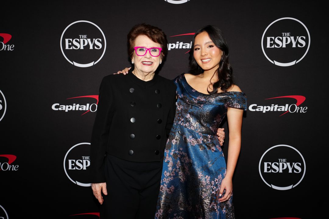 Two women pose together on a red carpet at the ESPYS, in front of a backdrop with ESPYS and Capital One logos. One wears a black outfit with bright pink glasses; the other, Julia Howe, shines in a blue patterned dress as she celebrates her biggest win with an ESPY Award.