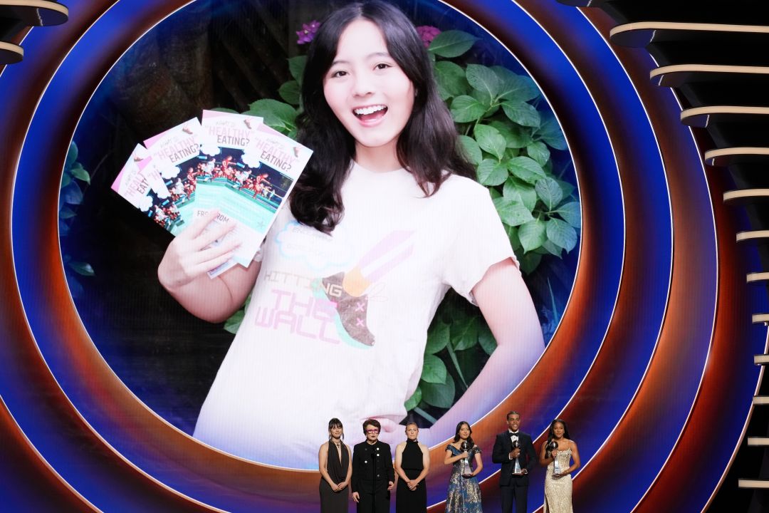 A large screen shows a smiling young woman, Julia Howe, holding several colorful flyers. On stage below, five people in formal wear—possibly presenting or accepting an ESPY Award—stand beneath the bright, concentric circles in the background.