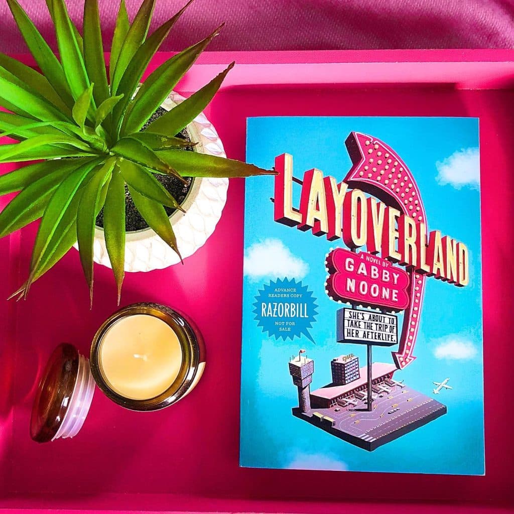 A pink tray holds a potted succulent, a lit candle in a glass jar, and the book Layoverland by Gabby Noone with a blue cover and a retro sign illustration—perfect inspiration for your next read while you listen to Gracie Abrams’ new album.