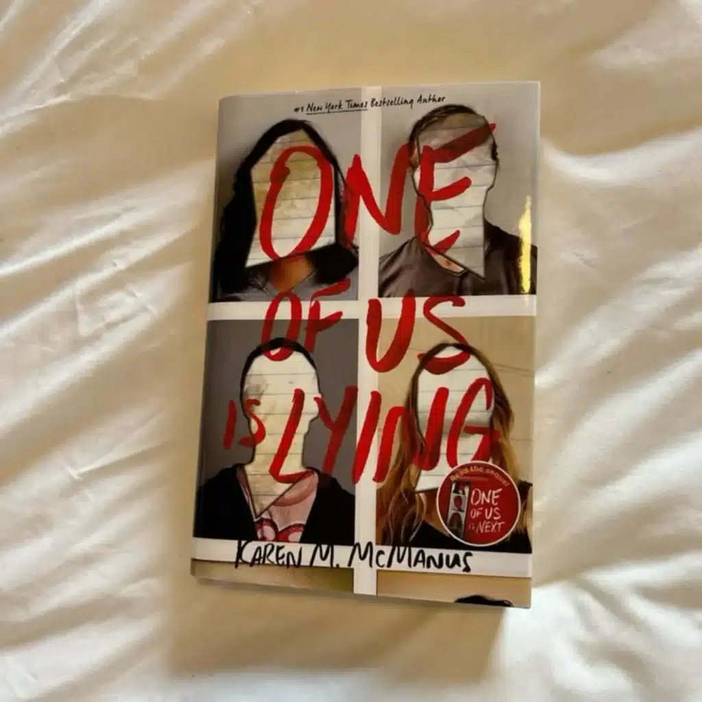 The book One of Us Is Lying by Karen M. McManus lies on a white bed. The cover shows four headshots with faces obscured, and the title is written in bold red letters across the images—making it the perfect next read to enjoy while listening to Gracie Abrams’ new album.