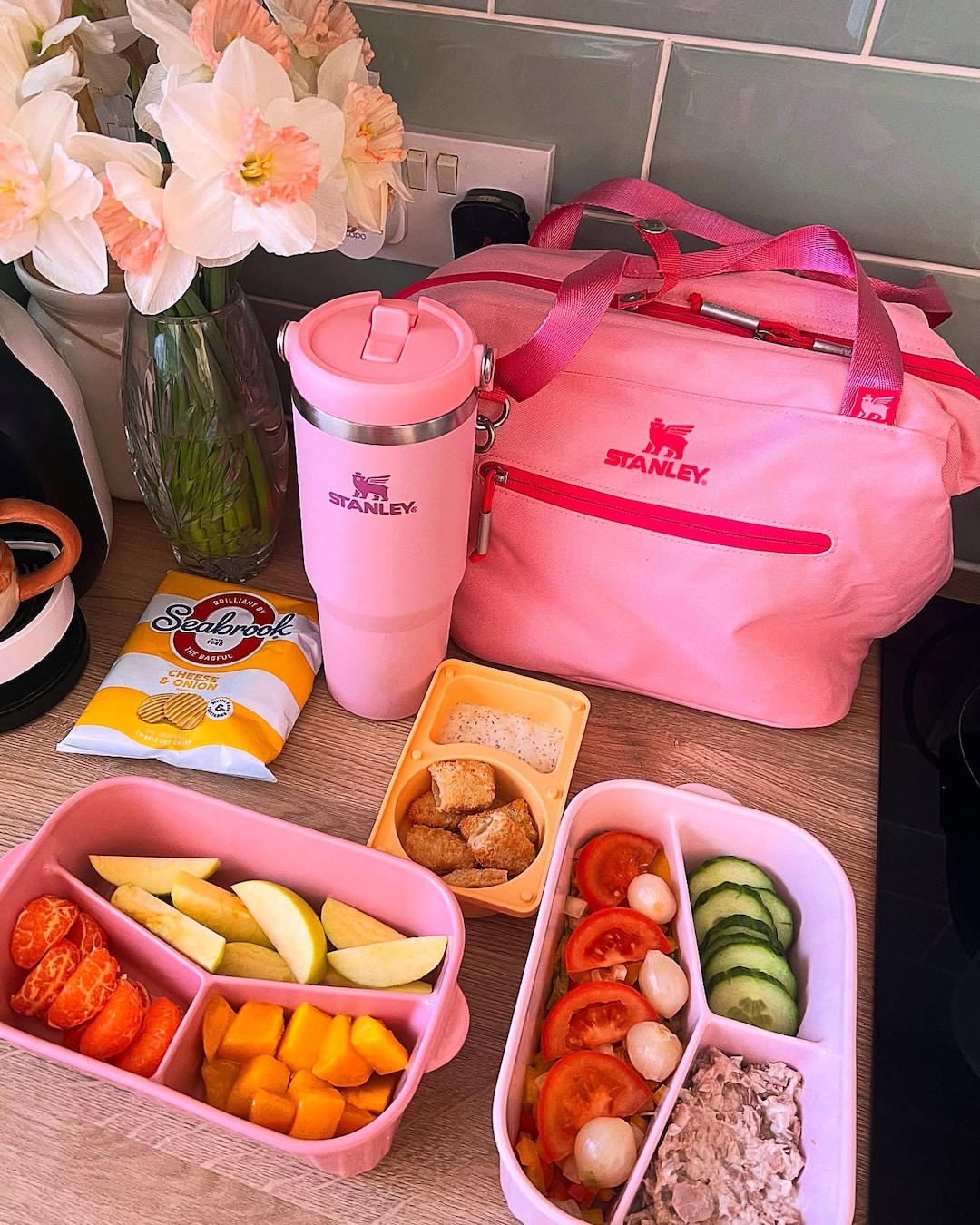 A pink lunch bag, tumbler, and bento boxes with fruit, vegetables, and tuna salad sit on a kitchen counter—an inviting scene of school preparation. Nearby are a flower vase, a kettle, and a bag of potato chips. The colorful and organized layout captures the thoughtful night-before-school routine.