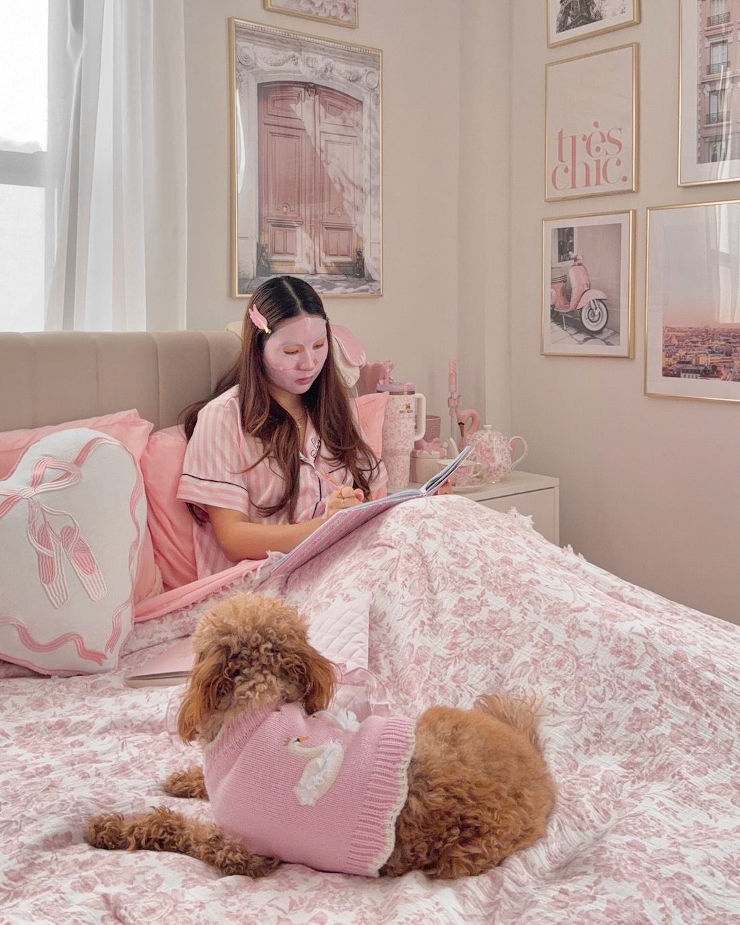 A woman in pink pajamas writes a to-do list in her notebook while sitting on a pink bed, preparing for the night-before-school. A brown dog in a pink sweater lies at the foot of the bed. The room is decorated with pink-themed art and ballet-inspired details.