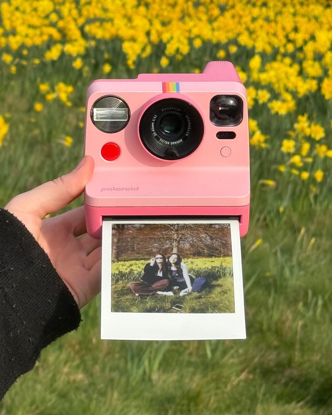 A hand holds a pink Polaroid camera outdoors with a photo printing out, showing two people sitting on grass. Yellow flowers fill the background, setting the perfect scene for a summer adventure where every snapshot feels like the start of a new side quest.