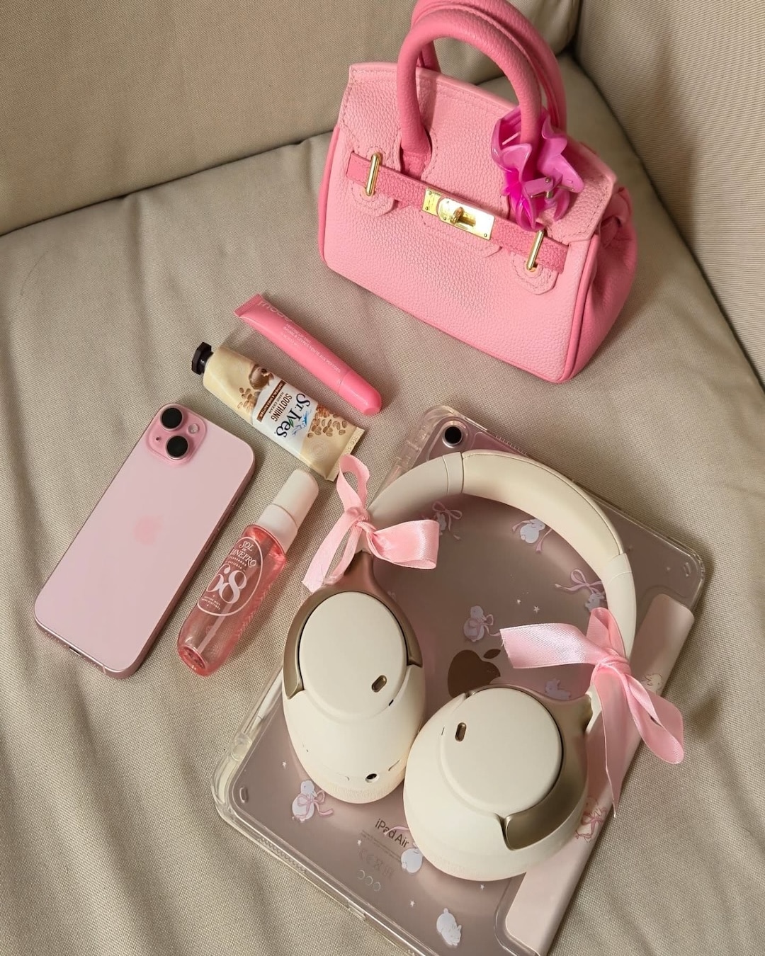 A pink handbag, pink iPhone, headphones with pink bows, lotion, lip gloss, spray bottle, and a tablet with a clear floral case are arranged on a beige surface—perfect essentials for your night-before-school to-do list and school preparation routine.