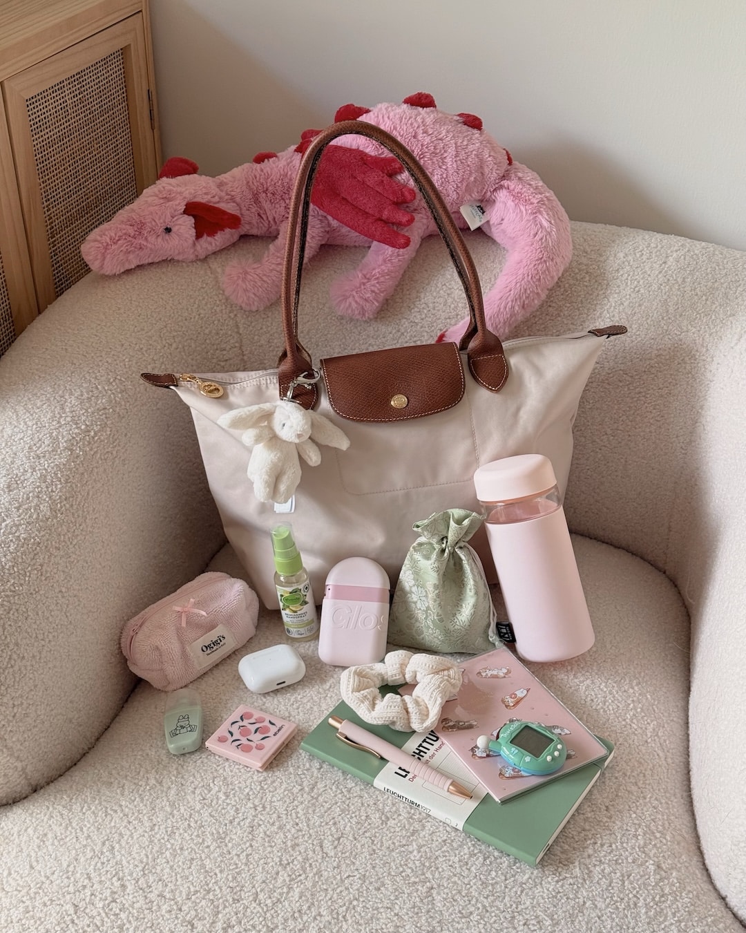 A beige bag on a cream chair with a pink plush dinosaur, pink water bottle, small pouch, notebook, hand cream, sanitizer, hairbrush, and soft baby accessories arranged neatly in front—perfect for your night-before-school school preparation or organizing your to-do list essentials.