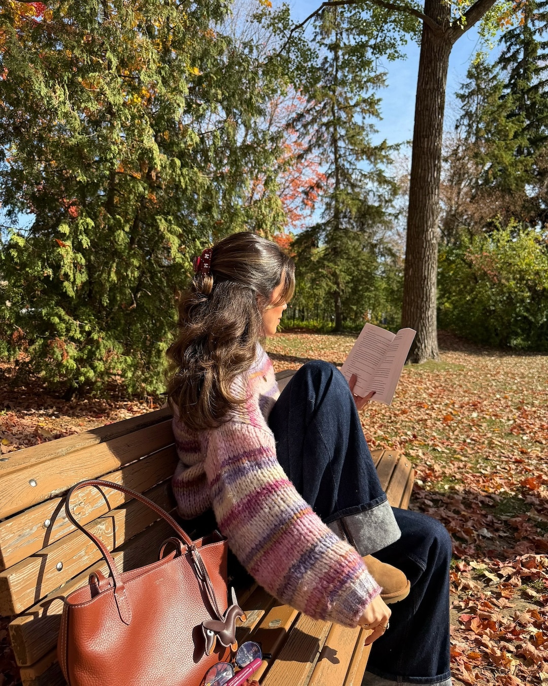 A person sits on a wooden bench outdoors, reading a book—perhaps a recent book rec from their favorite niche club. They wear a striped sweater and jeans, with a brown handbag resting beside them. Fallen leaves cover the ground, and trees with autumn foliage surround the scene, capturing the essence of another school year in full swing.