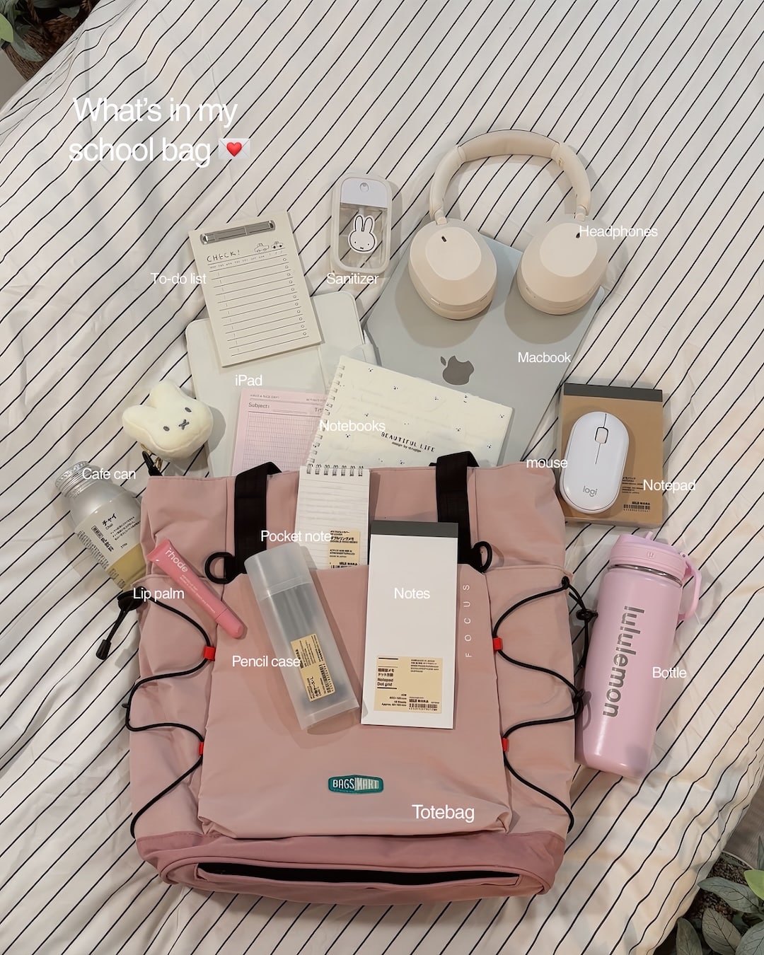 Pink tote bag on a bed with labeled items—MacBook, iPad, headphones, mouse, cat case, lip balm, pencil case, notes, notebooks, pocket notes, bottle, and stationery—arranged neatly around the bag as part of an organized night-before-school routine for easy school preparation.