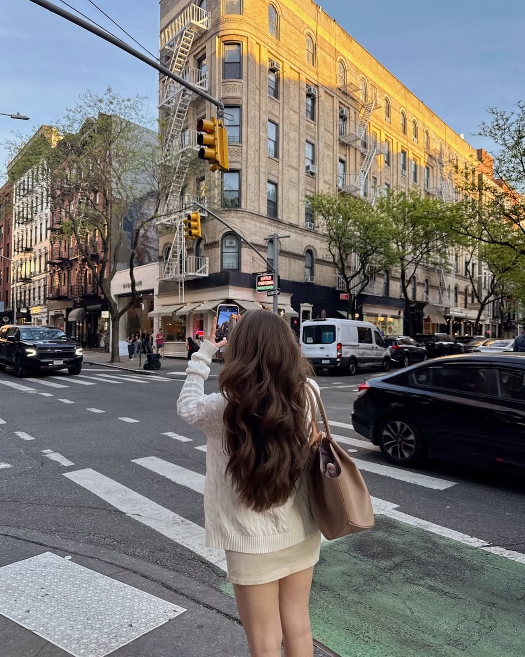A woman with long brown hair, dressed in a white outfit, stands at a city street corner taking a photo of a tan brick building under a blue sky, as if she’s the main character embarking on her own summer adventure. Cars and trees line the street, adding to the vibrant atmosphere of her urban journey.