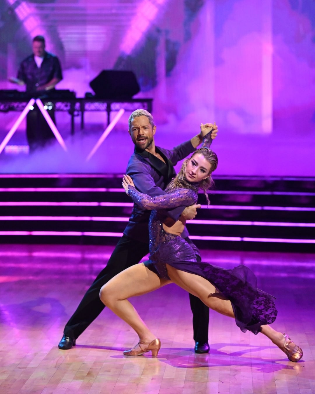 A man and woman in purple outfits perform a dramatic dance pose on a stage with colorful lighting, while a musician plays in the background—capturing the vibrant energy of Dancing With The Stars Week 2.