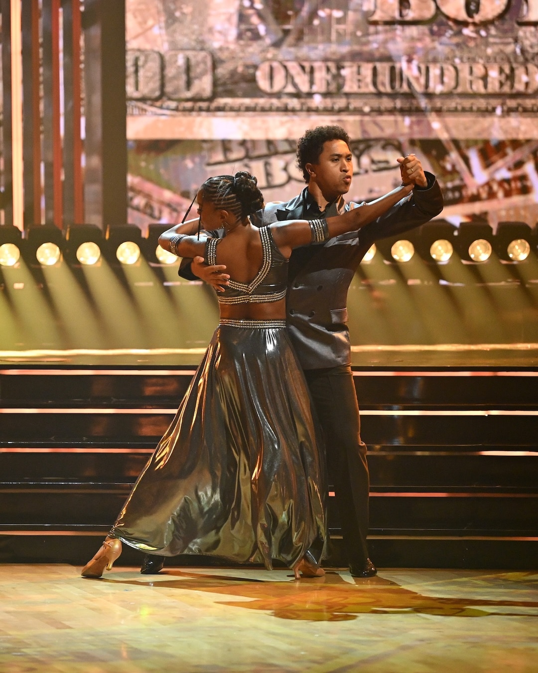 Two dancers in matching metallic outfits perform a ballroom dance on stage, with bright lights and a large dollar bill backdrop behind them, capturing the glitz and excitement of Dancing With The Stars during the Week 2 recap.