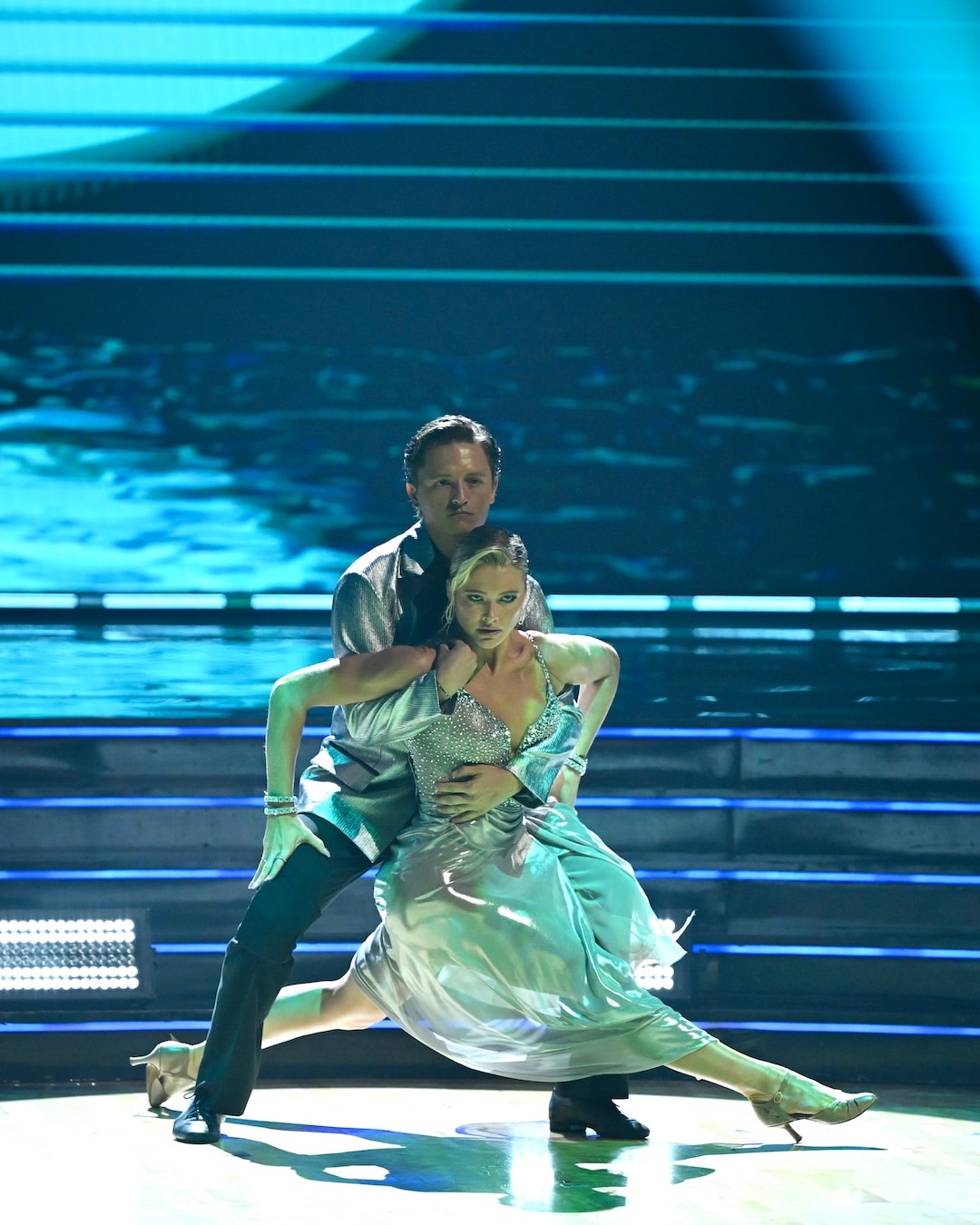 Two dancers perform a dramatic routine on stage. In this memorable Week 2 recap from Dancing With The Stars, the man stands behind the woman, holding her as she strikes a low pose, her dress shimmering under stage lights with illuminated stairs in the background.