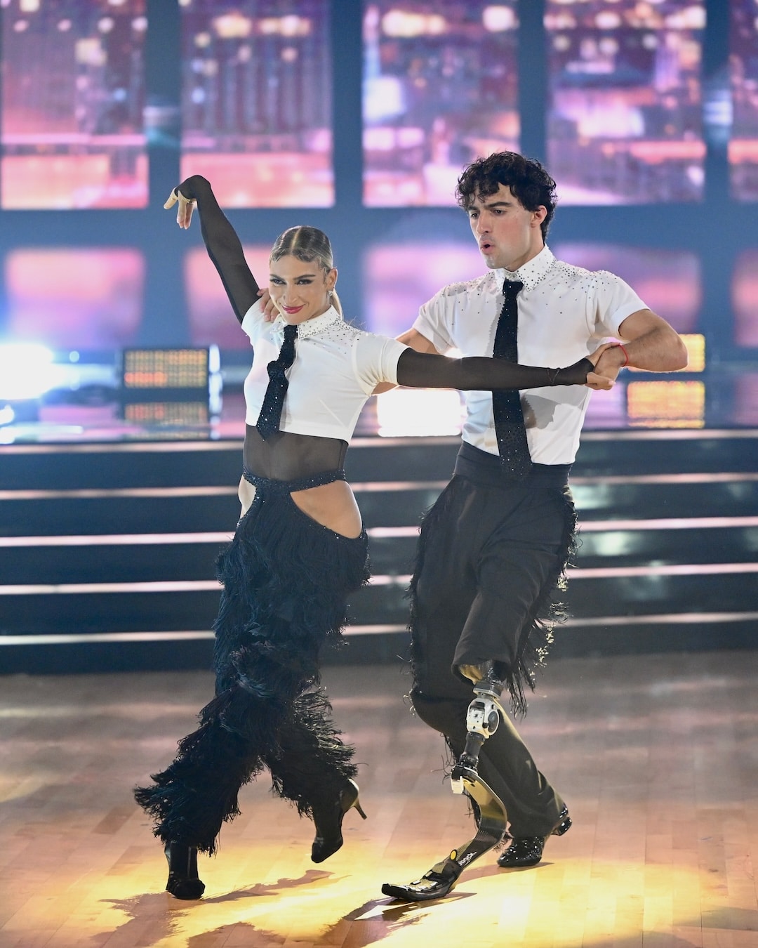 Two dancers in matching black and white costumes perform on a brightly lit stage in this exciting Week 2 recap of Dancing With The Stars. The male dancer has a prosthetic leg, and both have energetic expressions and dramatic poses, with sparkling lights and a blurred audience in the background.