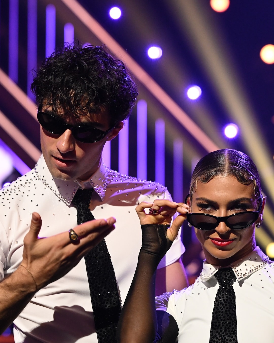 Two dancers in matching white shirts, black ties, and sunglasses pose confidently under stage lights; the woman lowers her glasses and smiles while the man gestures with his hand in a scene straight out of a Dancing With The Stars Week 2 Recap.