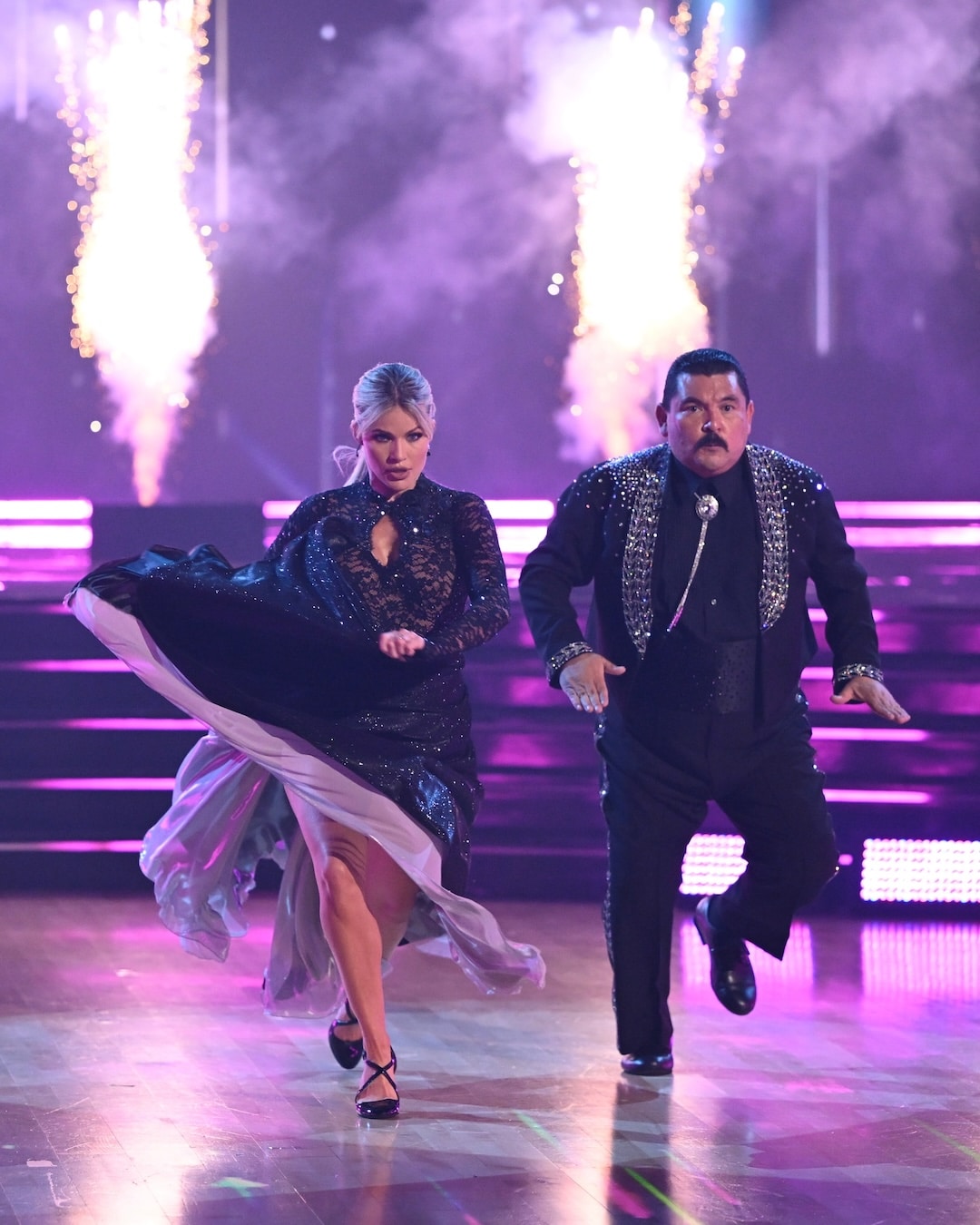 Two dancers in black outfits perform energetically on a lit stage with fireworks in the background. The woman lifts her dress while kicking, and the man dances beside her, both appearing focused and intense—a captivating moment from this week’s Dancing With The Stars Week 2 recap.