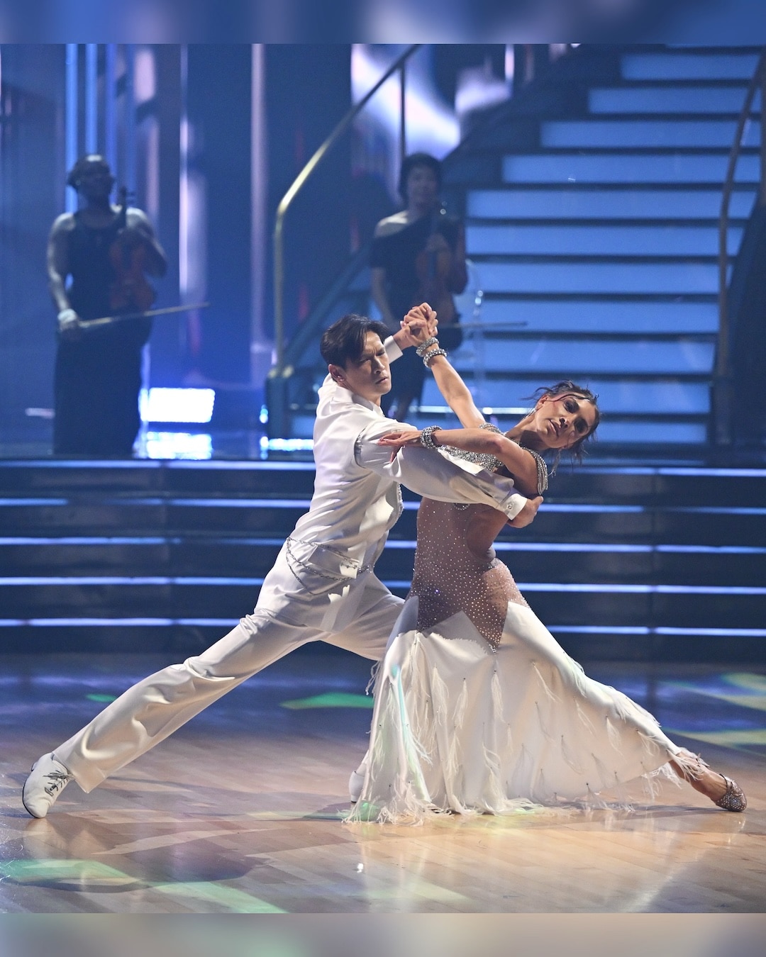 A couple in elegant dance attire performs a ballroom routine on a polished stage during week 2 of Dancing With The Stars, with stairs and musicians in the background under dramatic lighting.