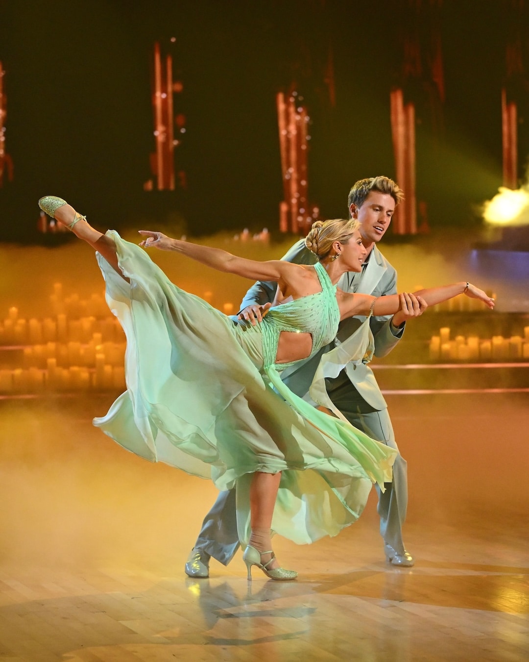 A man and woman in elegant mint green costumes perform a graceful ballroom dance pose on a wood floor, with the woman’s leg extended and sheer dress flowing, surrounded by soft stage lighting and mist—a captivating moment from Week 2 of Dancing With The Stars.