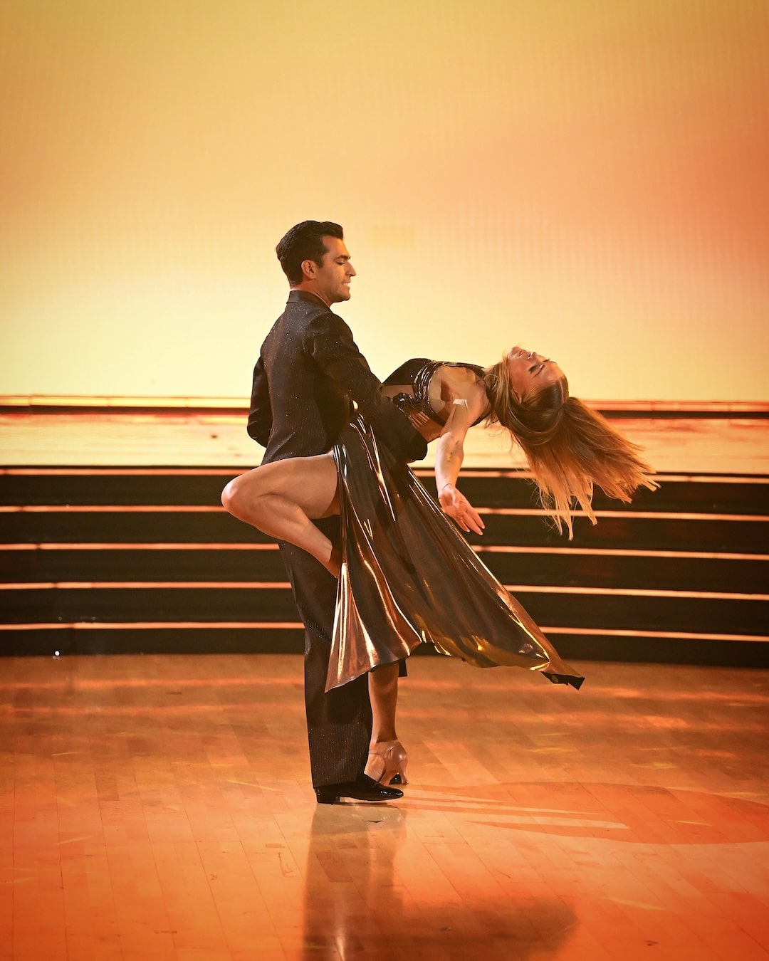 A man and woman perform a dramatic ballroom dance pose on a polished wooden floor, with the woman leaning back and her hair flowing, against a softly lit orange background—capturing the intensity of a Dancing With The Stars Week 2 recap.
