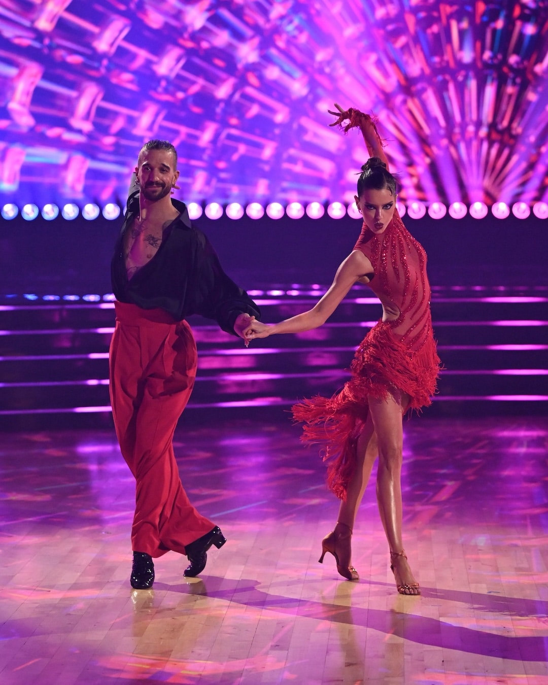 Two dancers perform on a brightly lit stage in this Week 2 recap of Dancing With The Stars. The man wears black and red, while the woman dazzles in a red fringe dress, striking a dramatic pose with one arm raised. The background glows with vibrant pink and purple lights.