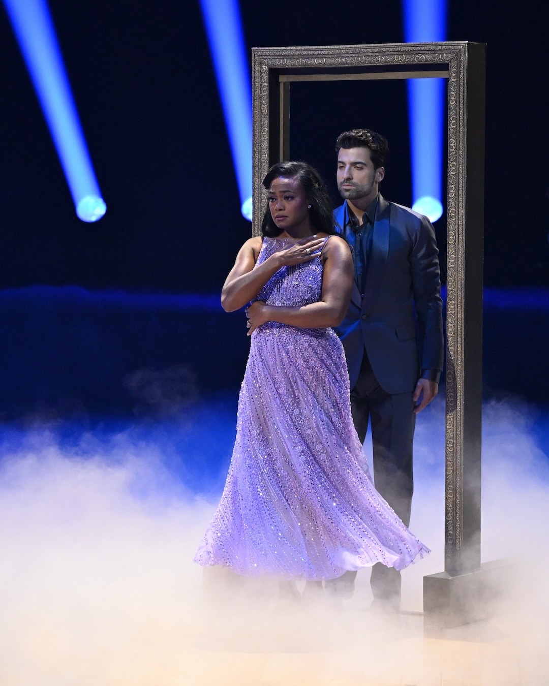 Two dancers stand closely together in a doorway frame on a foggy, dramatic stage for Dancing With The Stars, Week 2. The woman in front wears a sparkly lavender gown, while the man behind her wears a dark suit. Blue spotlights shine in the background.